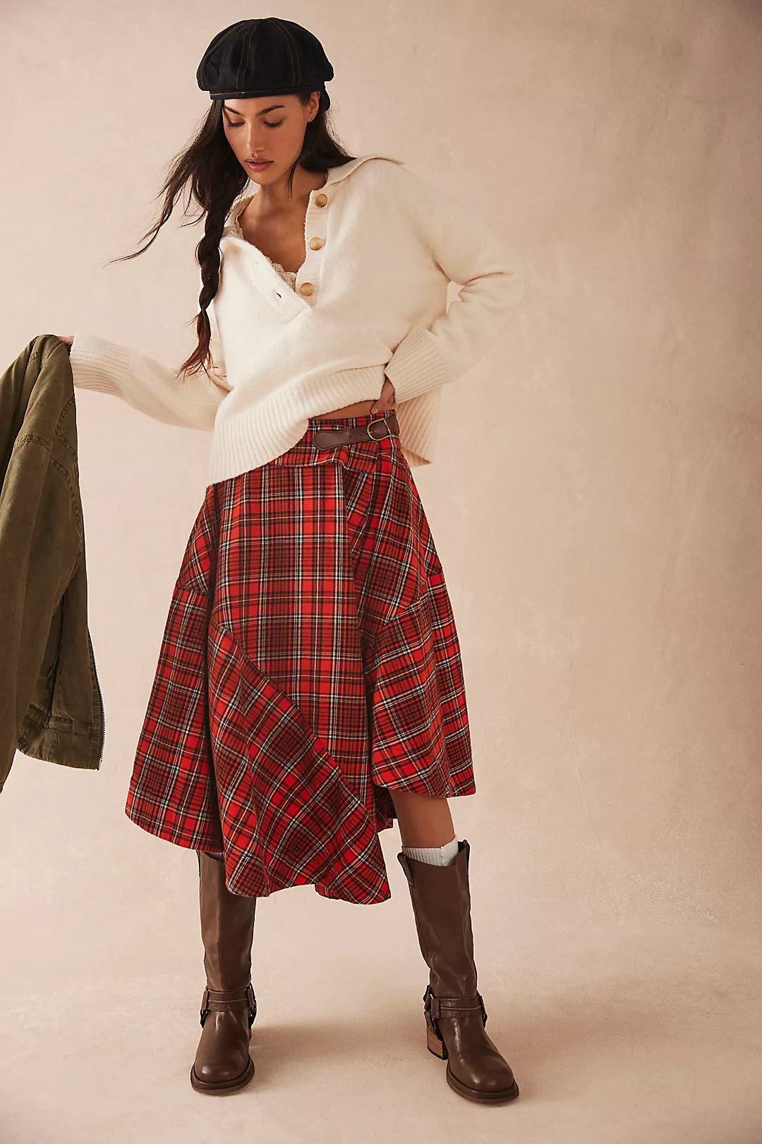 Woman wearing a white sweater, Free People red plaid skirt, and brown boots against a plain background
