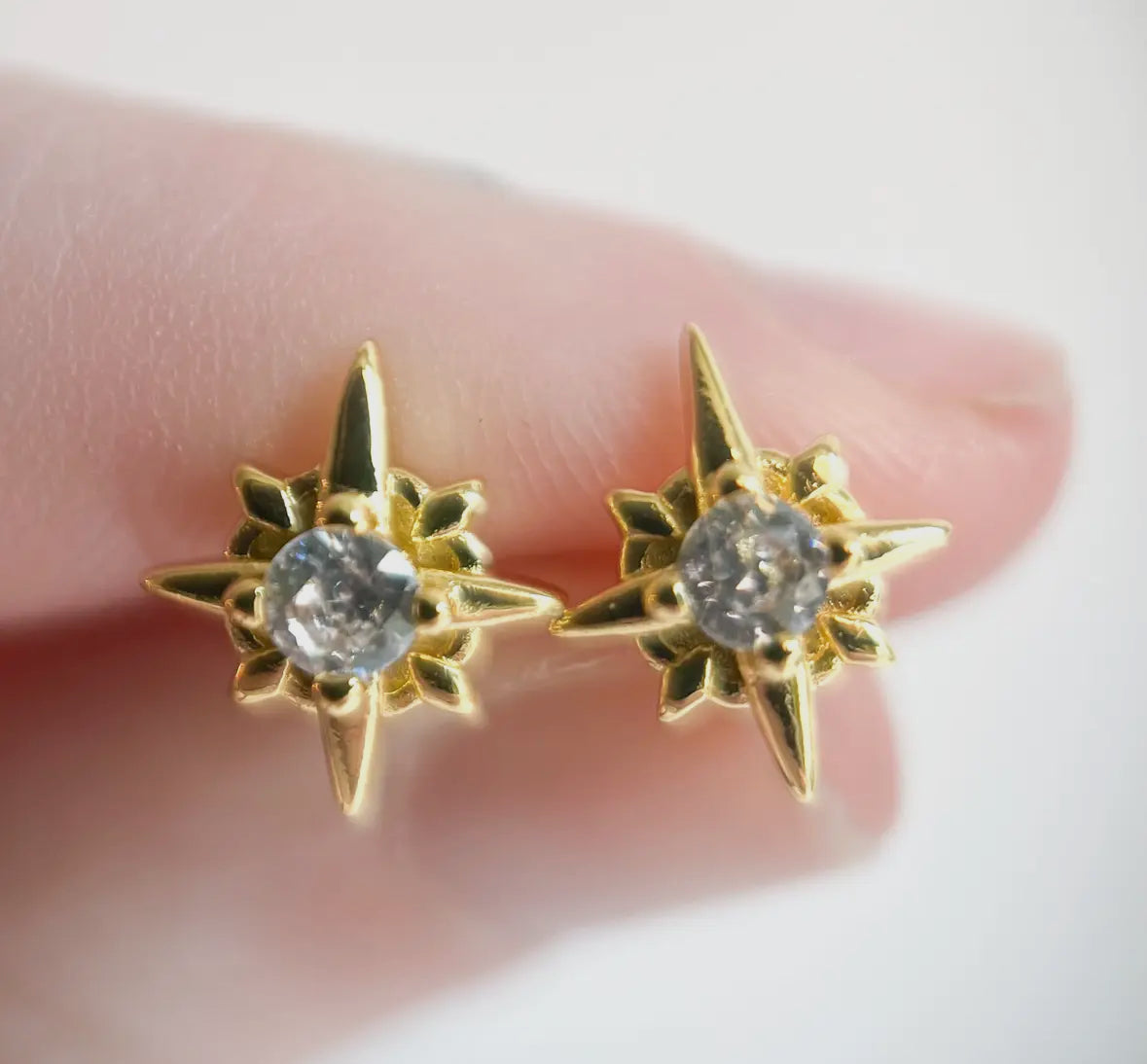 Gold star-shaped earrings with clear gemstones held between fingers on a white background