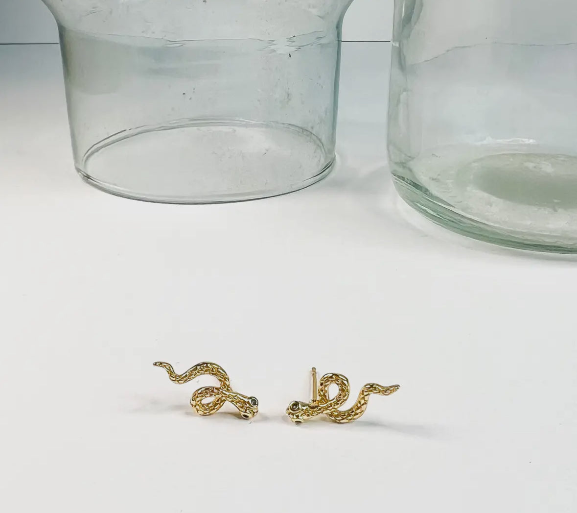 Gold snake-shaped earrings on a white surface with clear glass jars in the background.