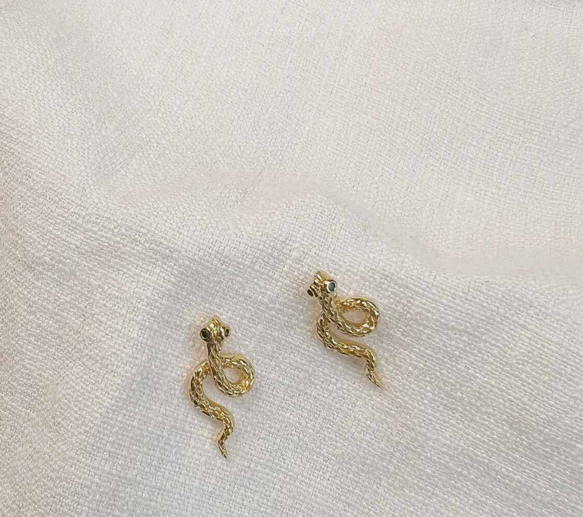 Gold snake-shaped earrings on a white fabric background