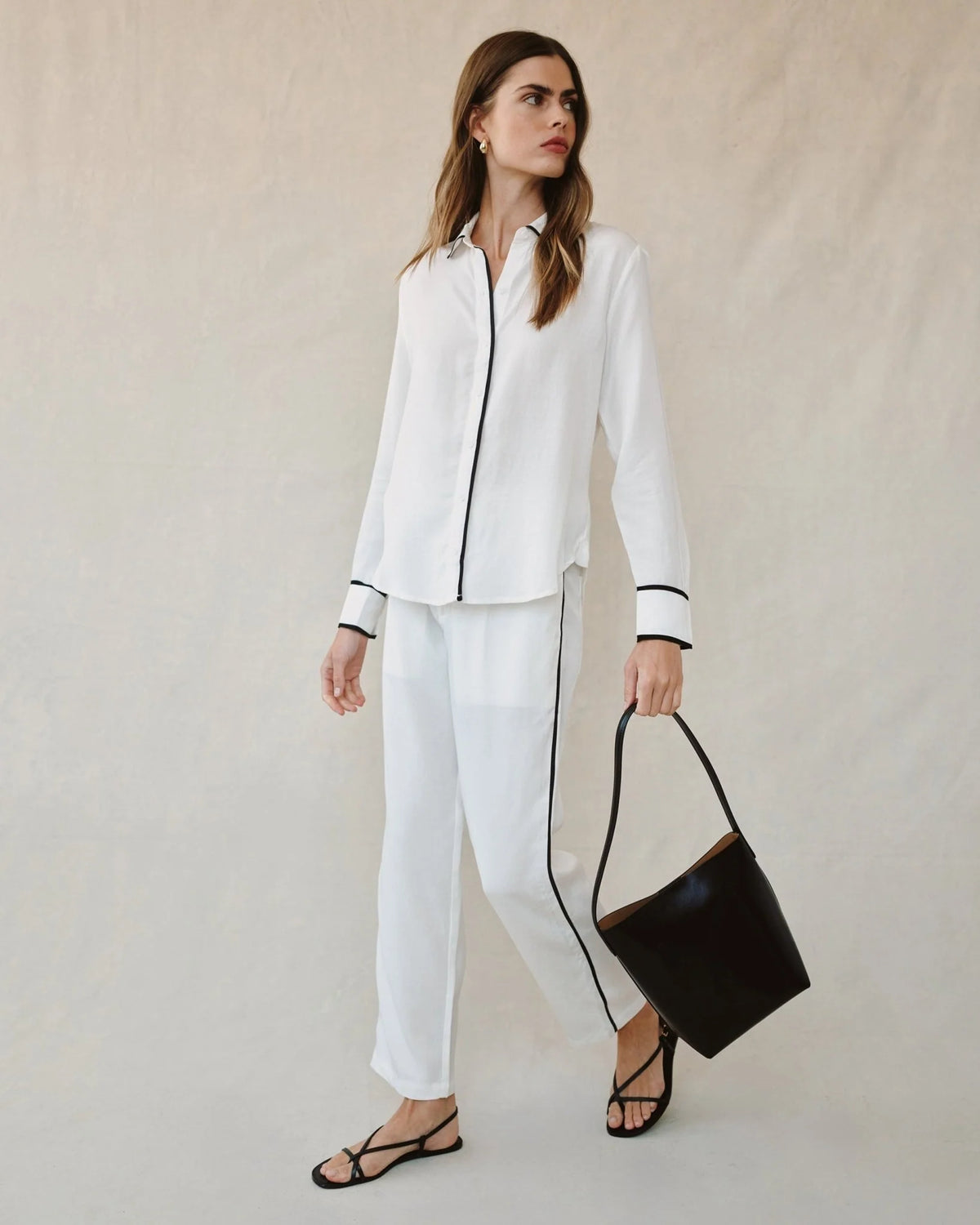 Woman wearing a white outfit with black accents and holding a black handbag on a plain background