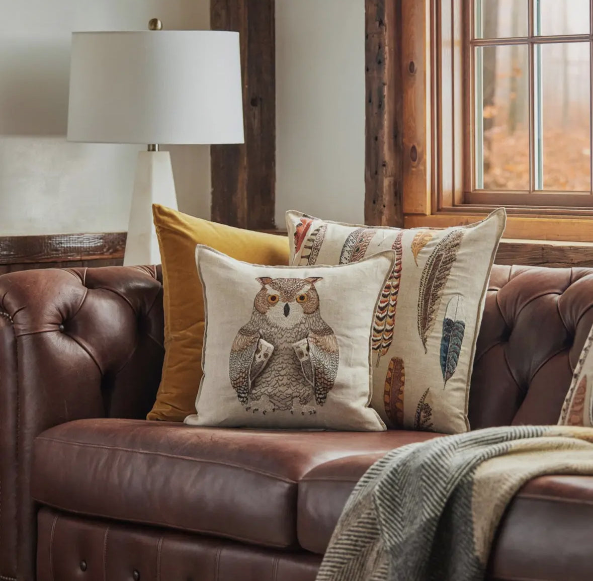 Brown leather sofa with decorative pillows featuring an owl design and feather patterns, in a cozy room setting.