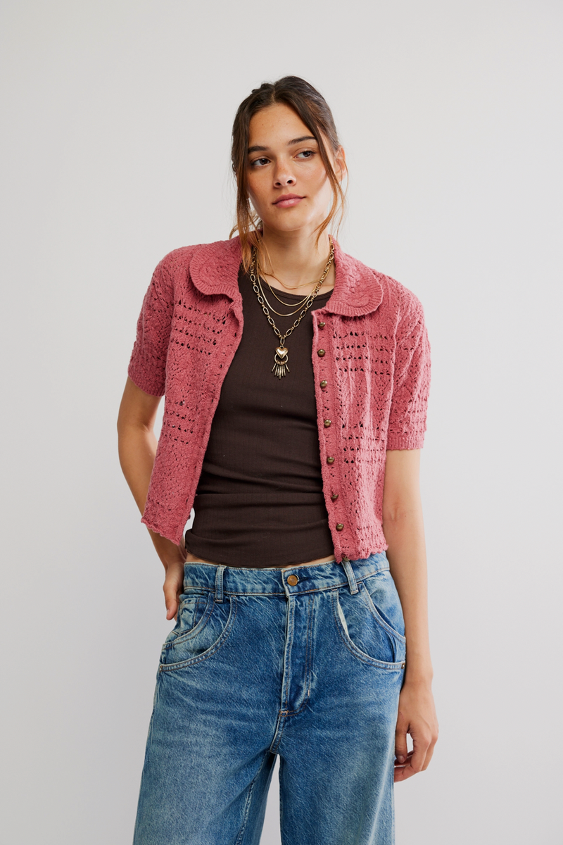 Woman wearing a pink textured cardigan, brown top, and blue jeans on a plain background