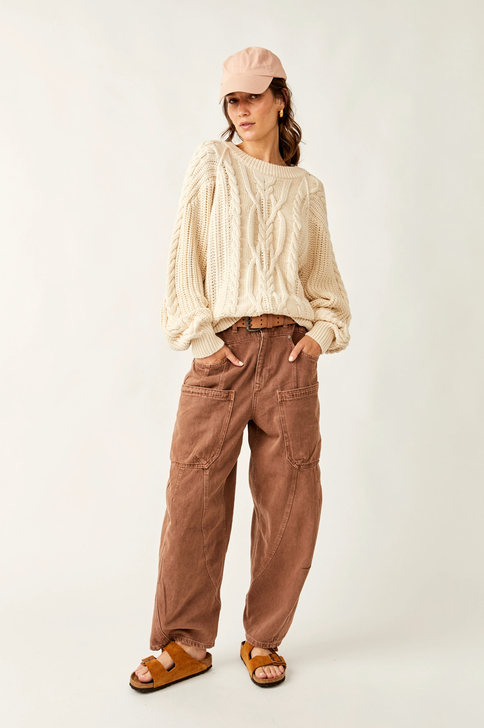 Person wearing a cream cable knit sweater, brown pants, and a pink cap on a white background
