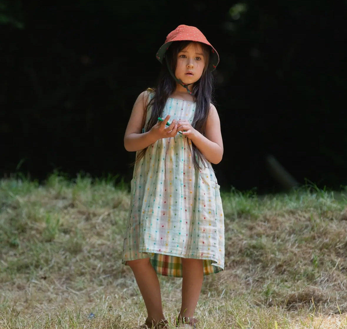 Young girl in a patterned dress and red hat standing in a grassy field with a dark background