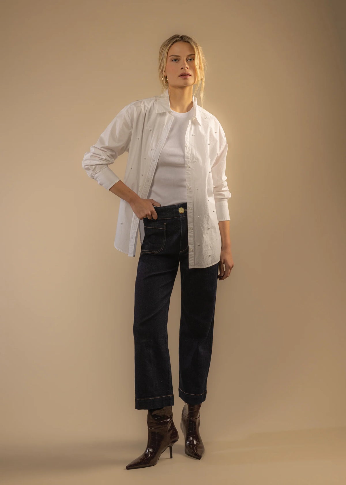 Woman wearing a white shirt, dark jeans, and brown shoes against a beige background