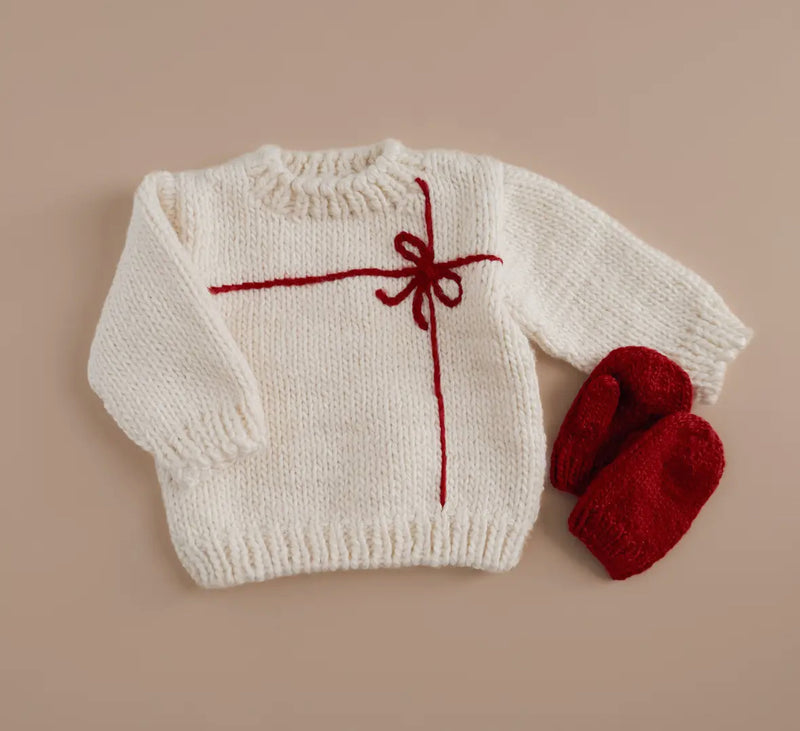 White baby sweater with red bow design from Blueberry Hill and matching red mittens on a beige background.