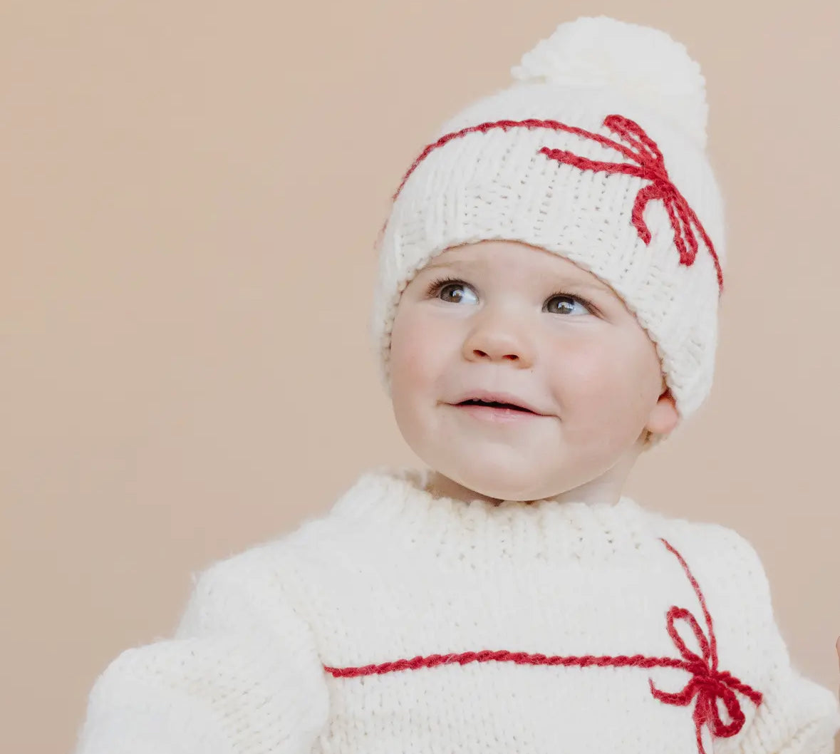 Baby wearing a white knit outfit with red bows from Blueberry Hill against a beige background. 