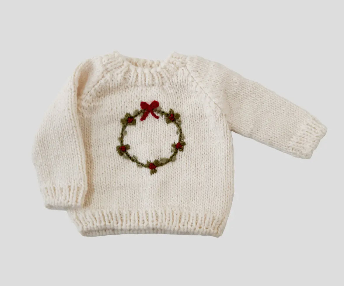 White knitted baby sweater with a Christmas wreath design on a light gray background.