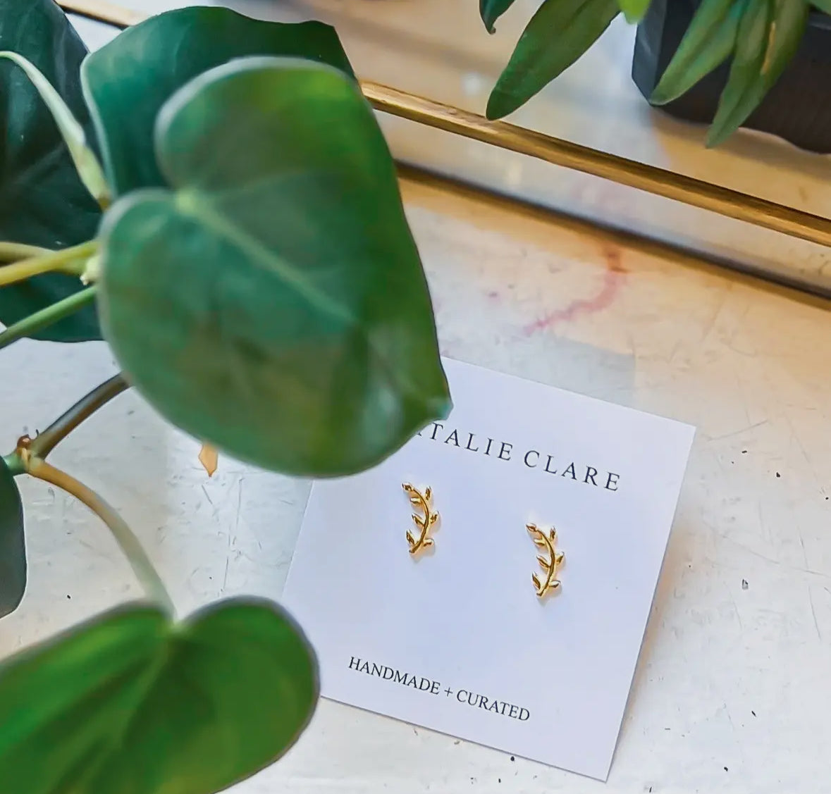 Gold earrings on a 'Natalie Clare' card with green leaves in the foreground