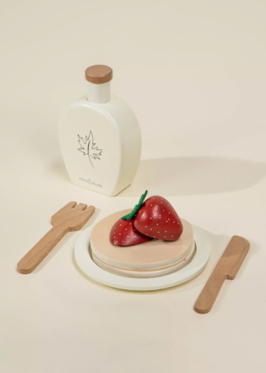 White ceramic bottle with wooden cap, wooden fork, knife, and small round pink cake with red decorations on a beige background.