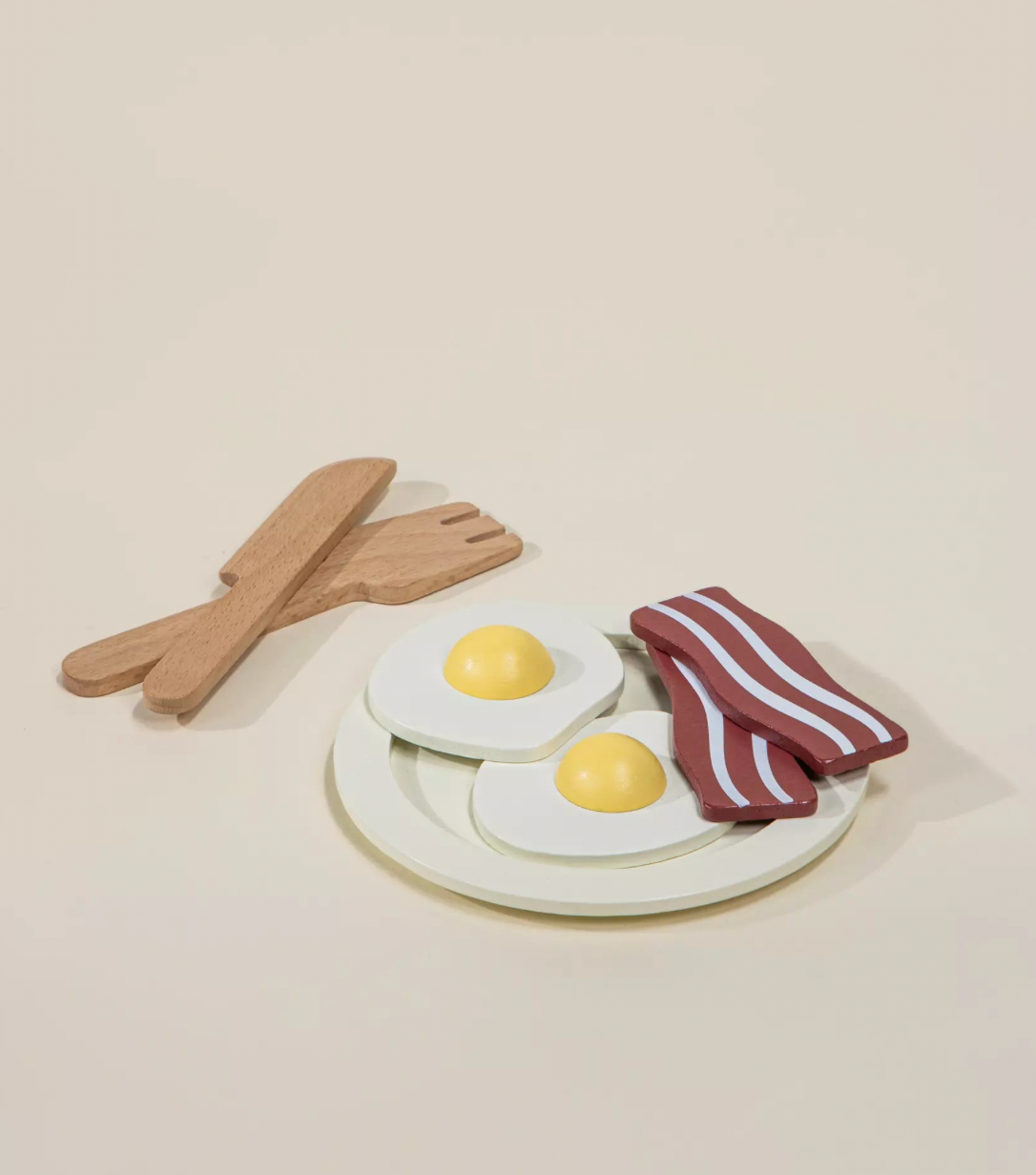 Toy set resembling a plate with eggs and bacon, accompanied by wooden utensils on a beige background.
