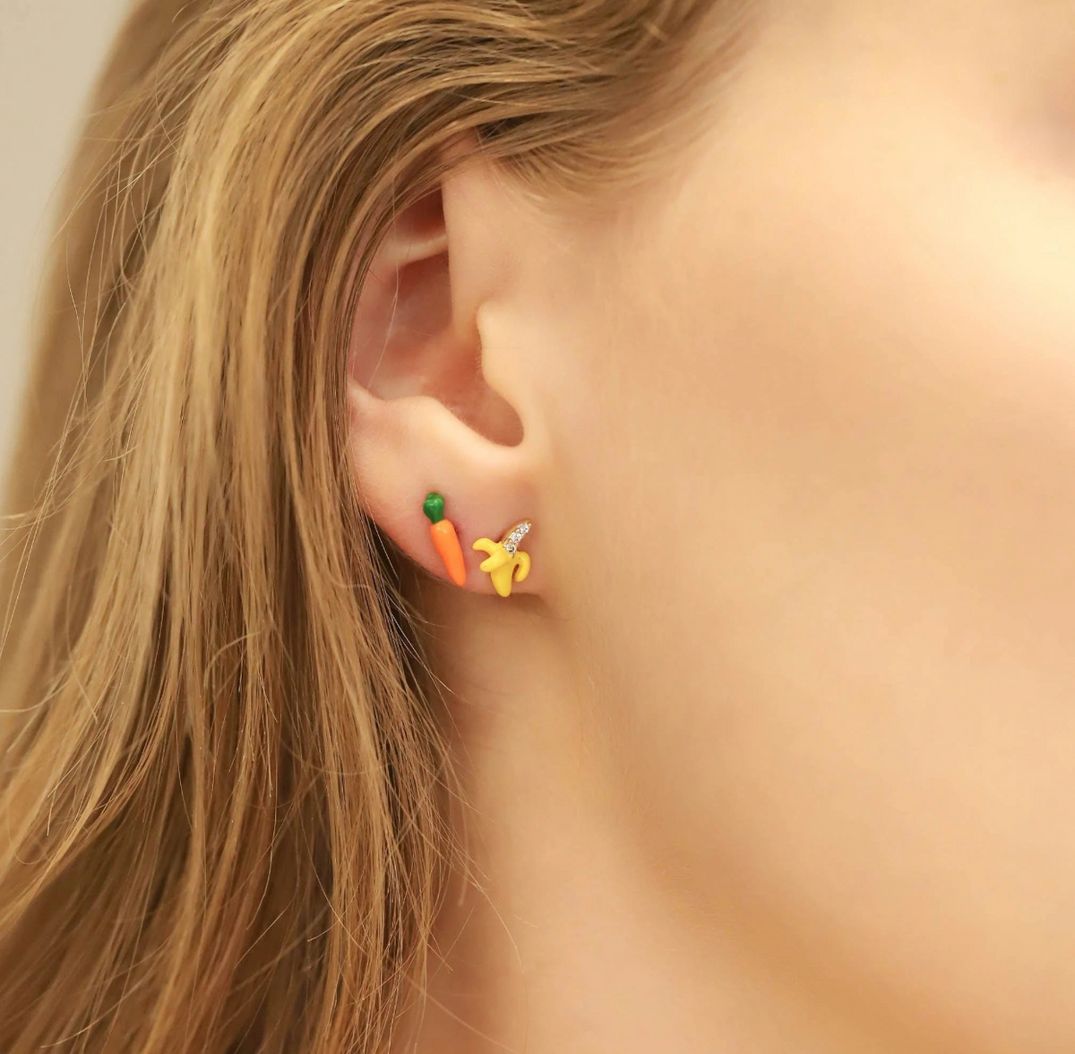 Close-up of an ear wearing a carrot-shaped earring with a banana-shaped earring.