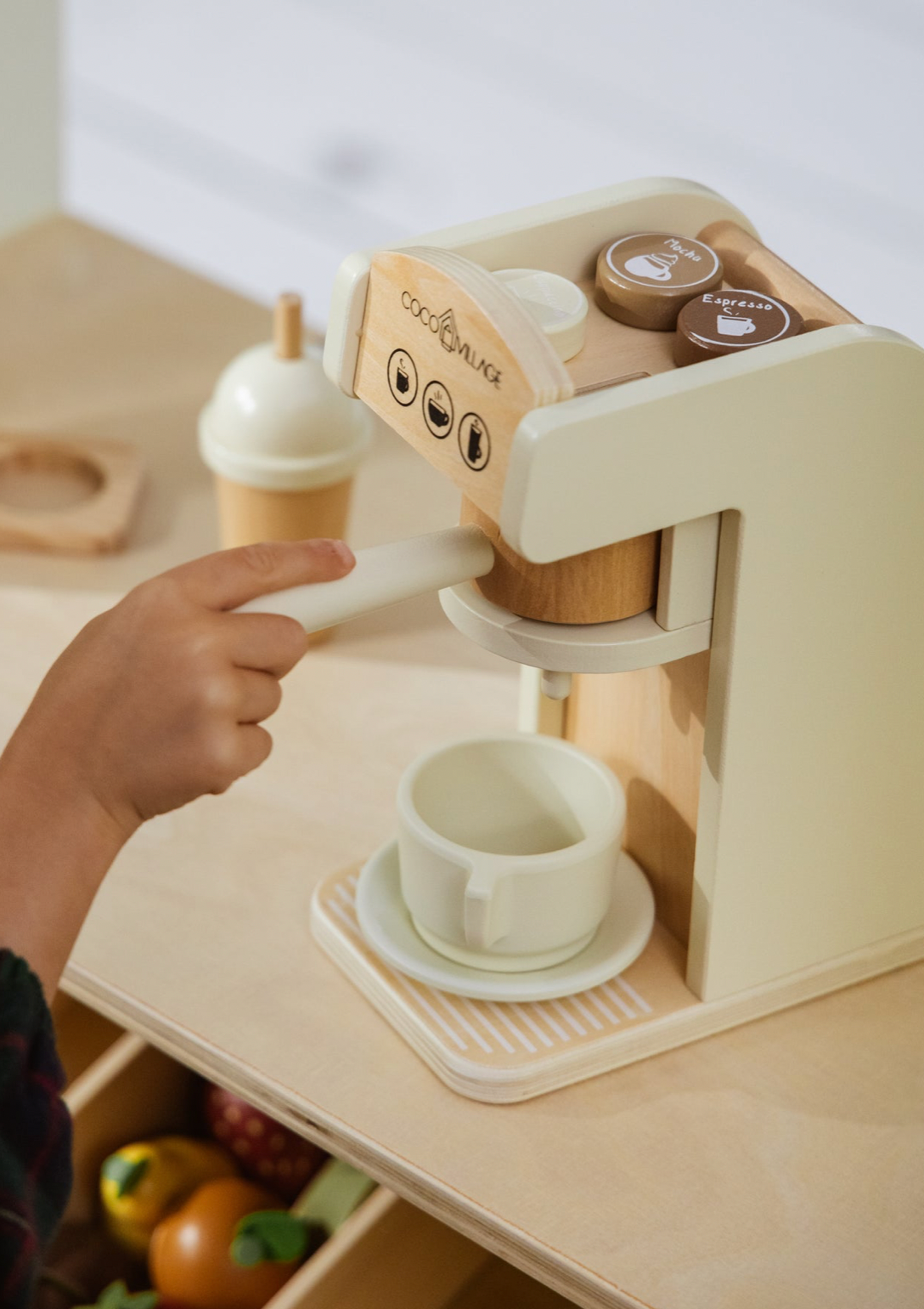 Wooden toy coffee machine with a child's hand pouring a cup, on a light wooden surface.