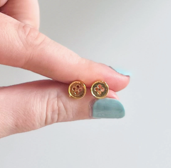 Gold button-shaped earrings held between fingers against a neutral background