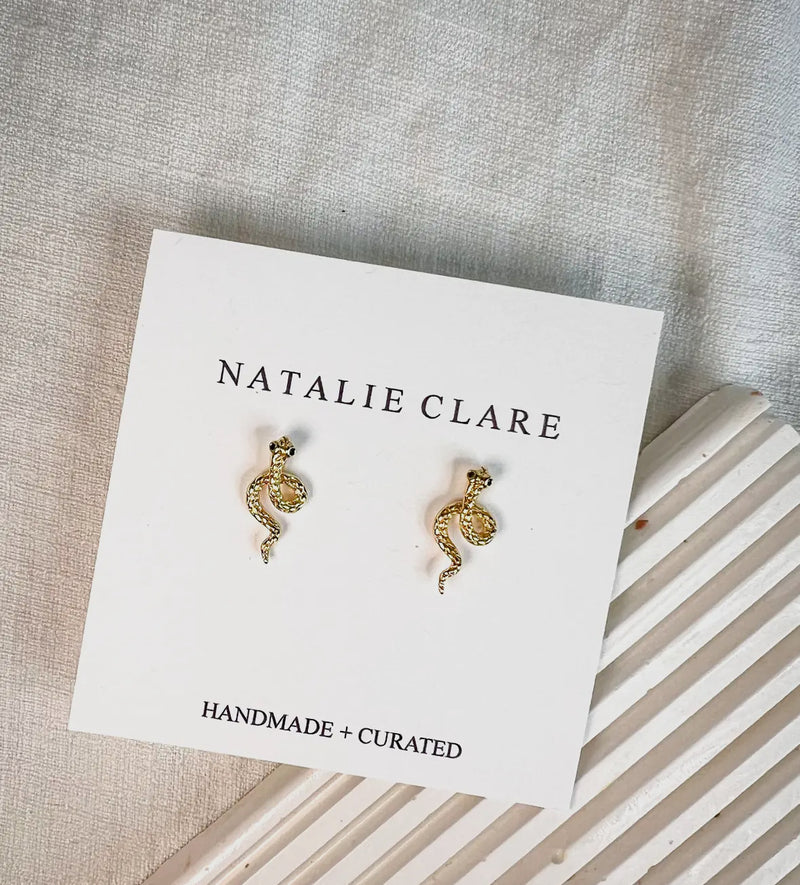 Gold snake-shaped earrings on a 'Natalie Clare' branded card with a striped surface underneath.