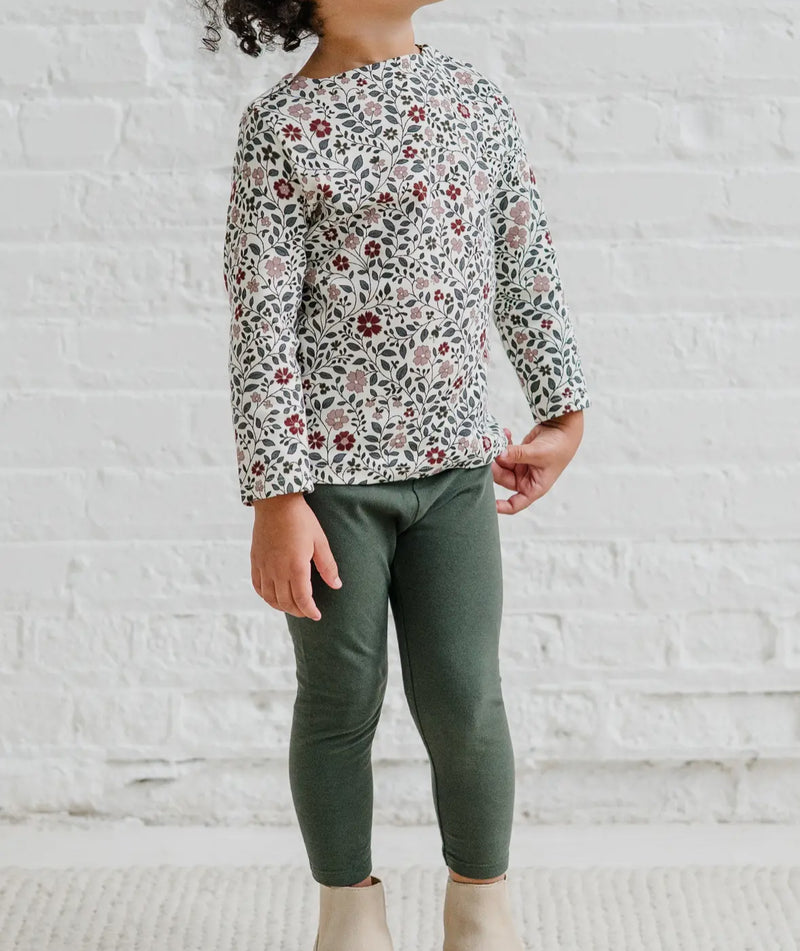 Child wearing a floral long-sleeve shirt and green pants against a white brick wall.