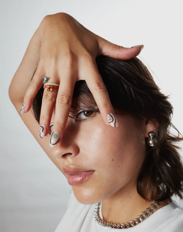 Person with hand on face, wearing jewelry including rings and earrings, against a plain background