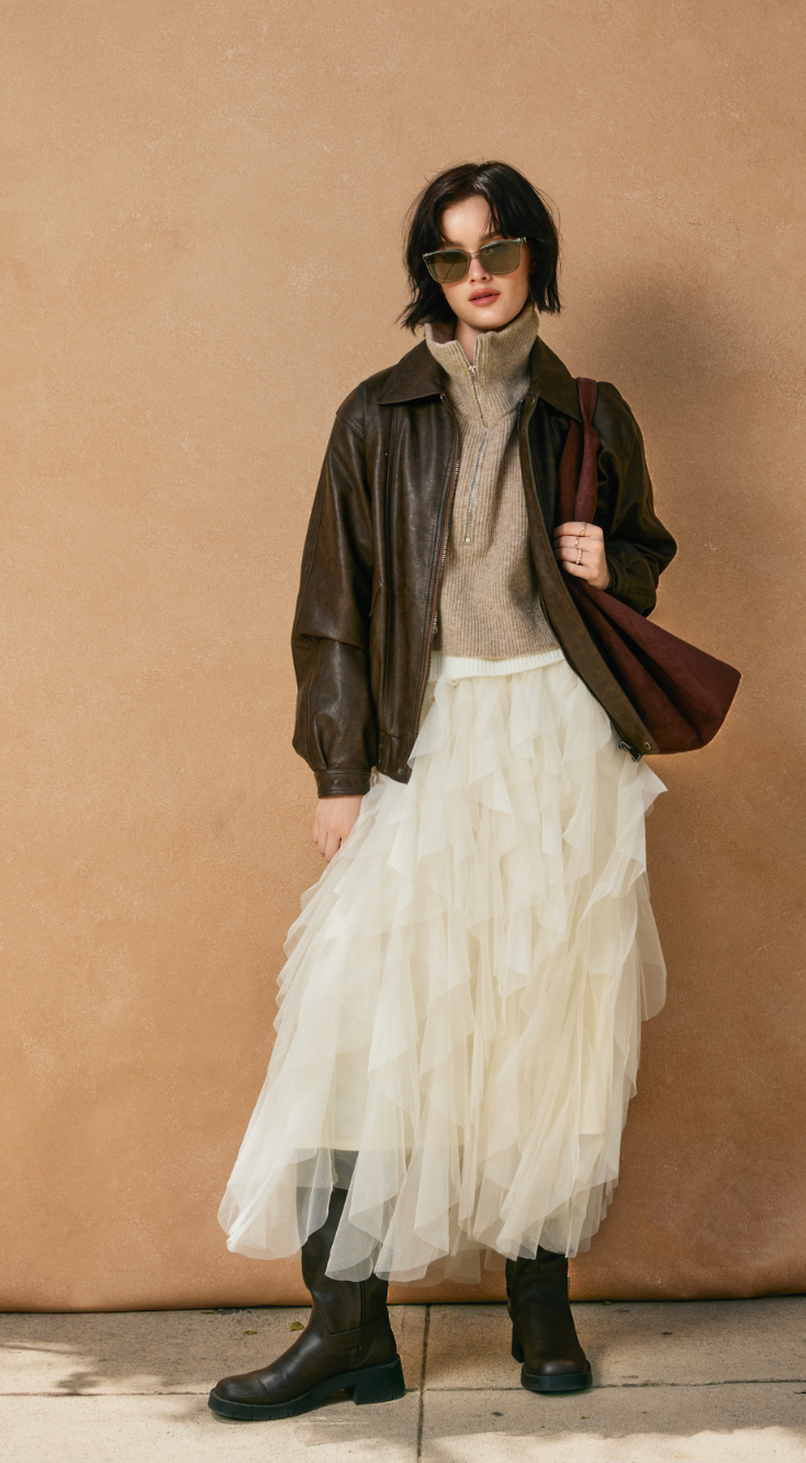 Person wearing a brown leather jacket, beige sweater, white tulle skirt, and black boots against a beige wall.