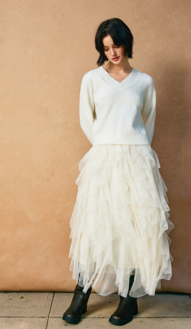Woman wearing a white sweater and layered white skirt against a beige wall.
