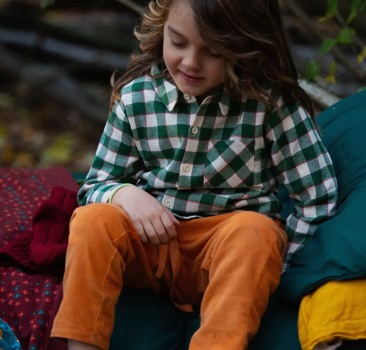 Child wearing a green and white checkered shirt and orange pants sitting outdoors.