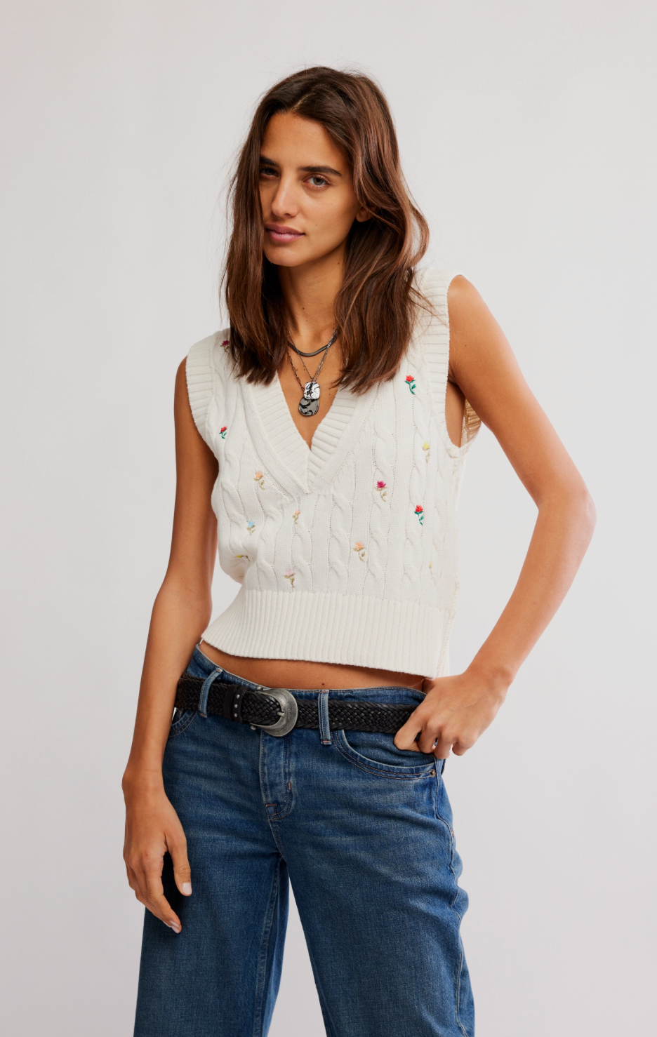 Woman wearing a white sleeveless sweater with colorful buttons and blue jeans on a plain background