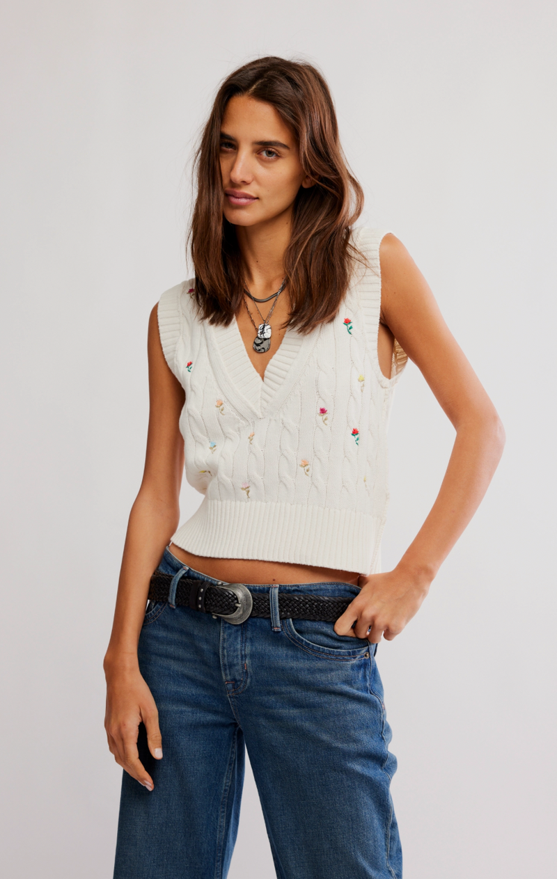 Woman wearing a white sleeveless sweater with colorful buttons and blue jeans on a plain background