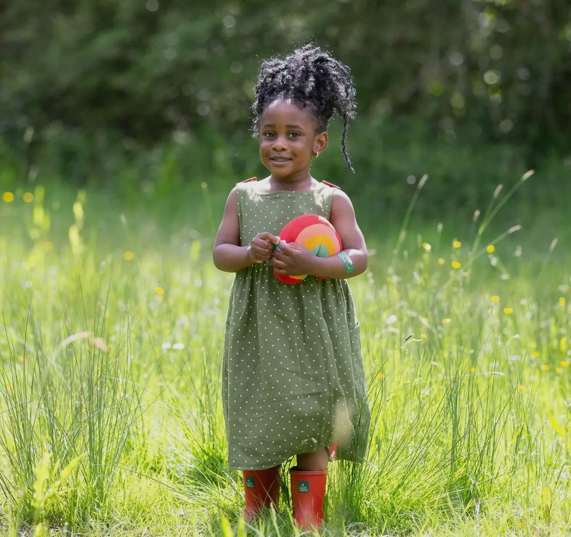 Young girl in a green dress and red rain boots standing in a grassy field holding a colorful ball.