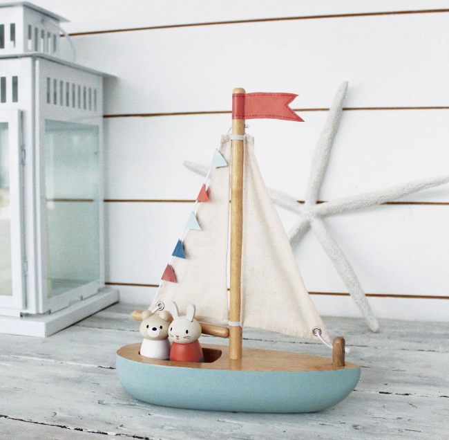 Toy sailboat with a white sail and red flag on a wooden surface, with a white lantern and windmill in the background.