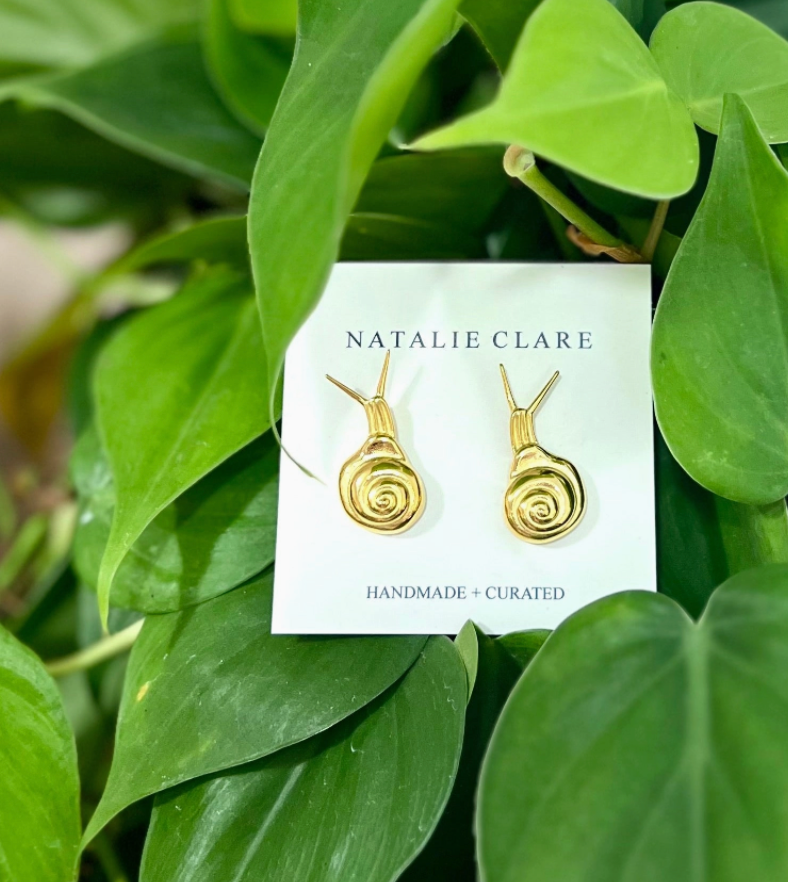 Gold snail-shaped earrings on a 'Natalie Clare' card with green leaves in the background