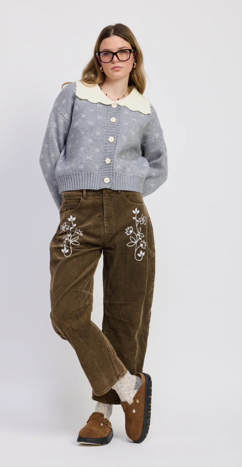 Woman wearing a gray cardigan, white shirt, brown pants with floral embroidery, and brown shoes on a white background