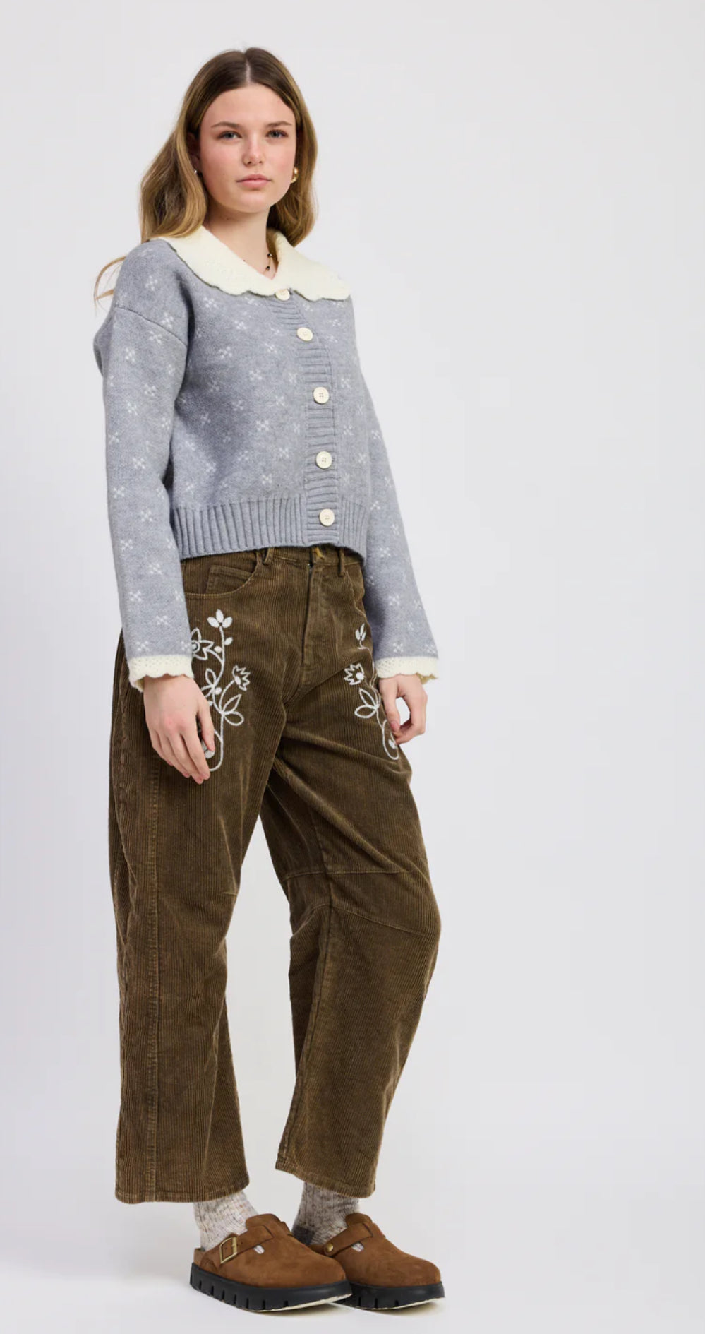 Woman wearing a gray sweater with white buttons and brown pants on a white background