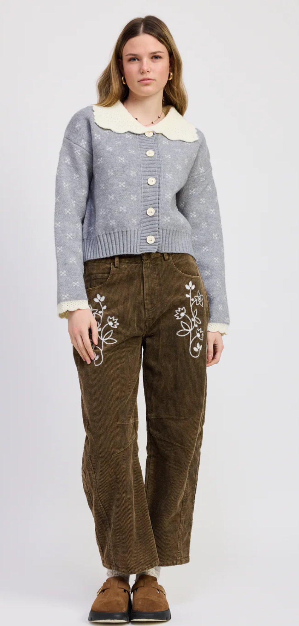 Woman wearing a gray patterned cardigan and brown pants with floral embroidery on a white background