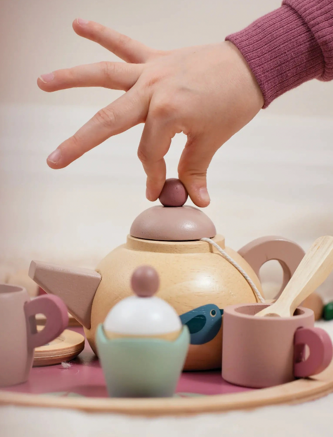 Child's hand interacting with a toy tea set on a light surface.