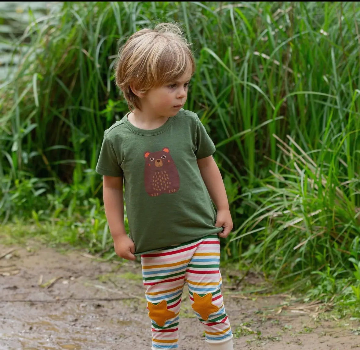 Child wearing a green t-shirt with a bear design and colorful pants standing in a natural setting.