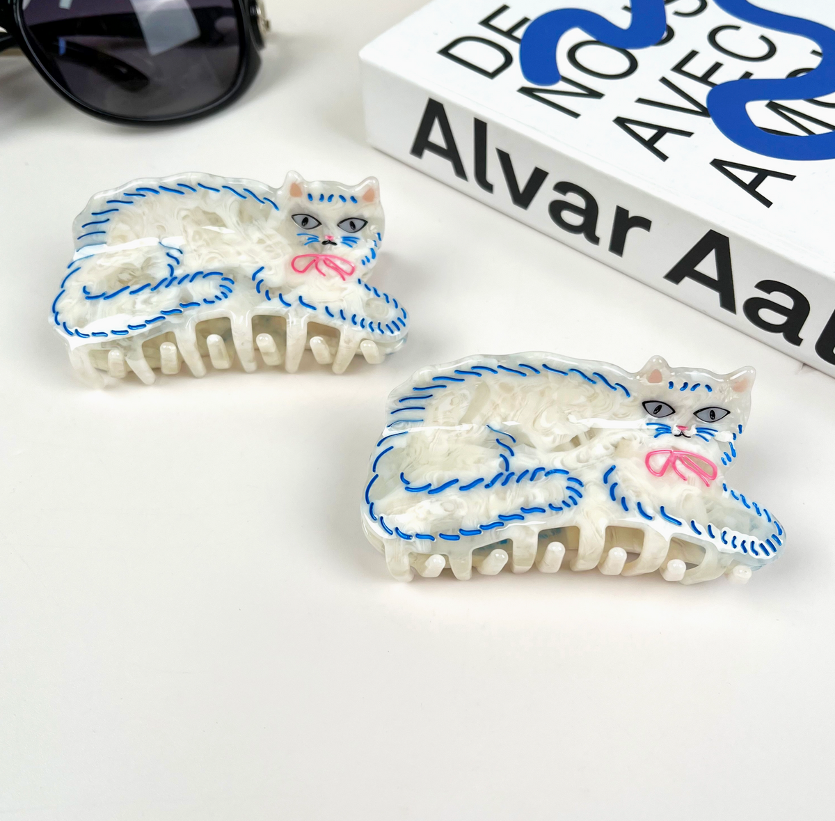 Two cat-shaped hair clips on a white surface with sunglasses and a book in the background.