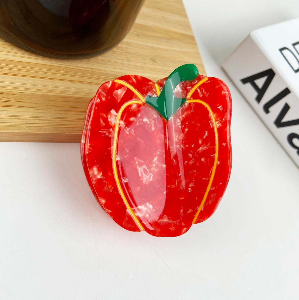 Red pepper-shaped object on a white surface with a wooden board and white box in the background.