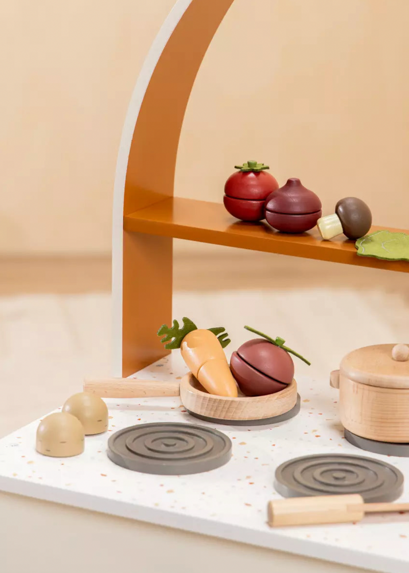 Wooden toy kitchen set with pots, pans, and vegetables on a beige surface.