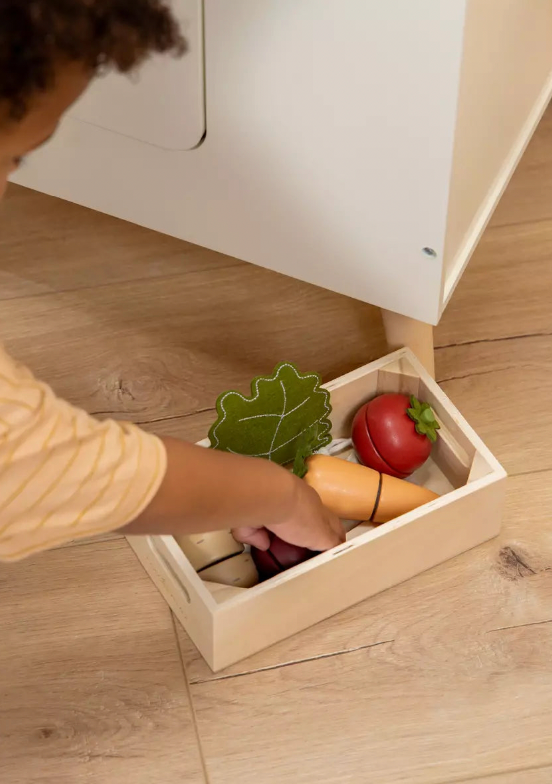 Child playing with toy fruits and vegetables in a wooden box on a wooden floor.