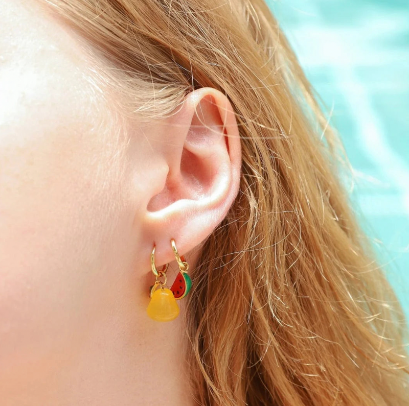 Close-up of an ear wearing a small hoop earring with a yellow charm and red heart charm, against a blurred background.
