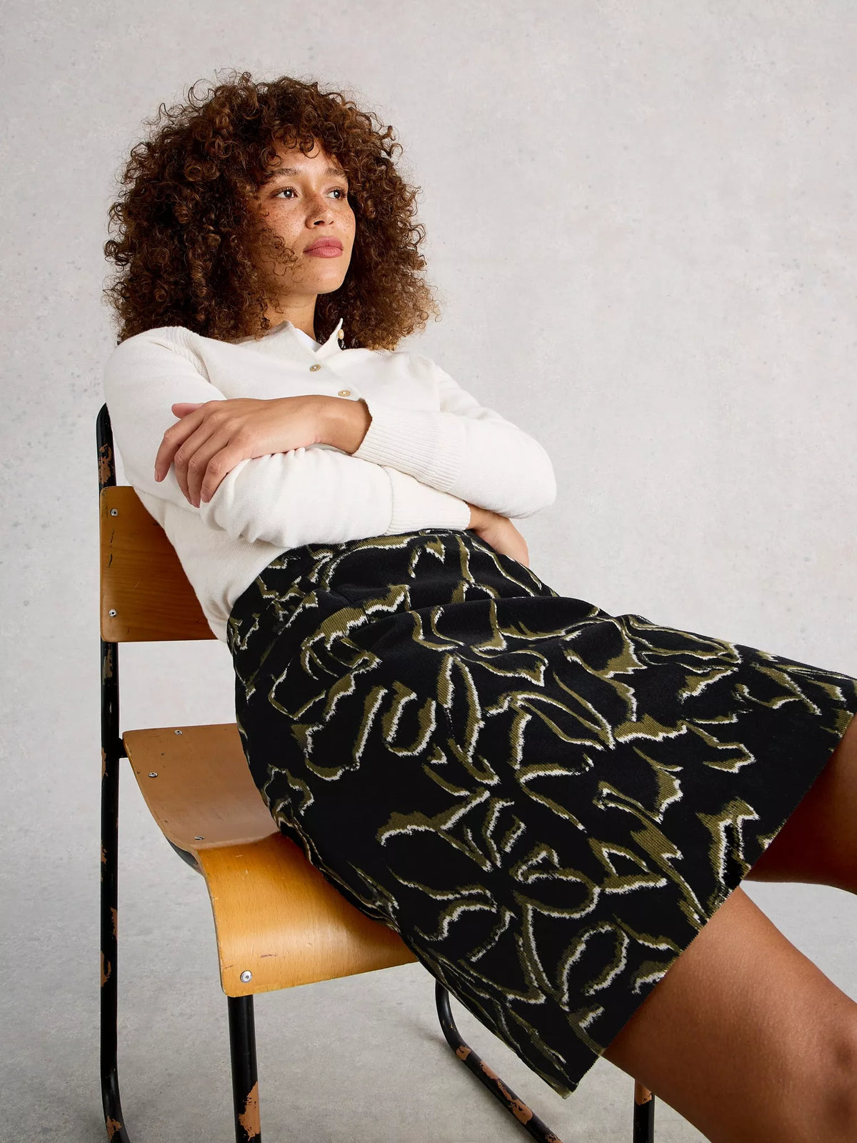 Woman sitting on a chair wearing a white blouse and black patterned skirt.