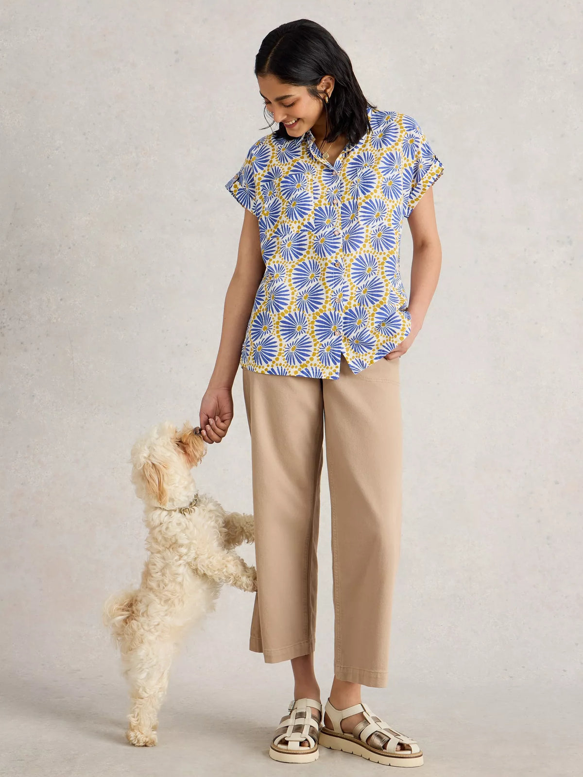 Woman wearing a blue and yellow patterned shirt and beige pants, standing next to a small dog on a plain background.