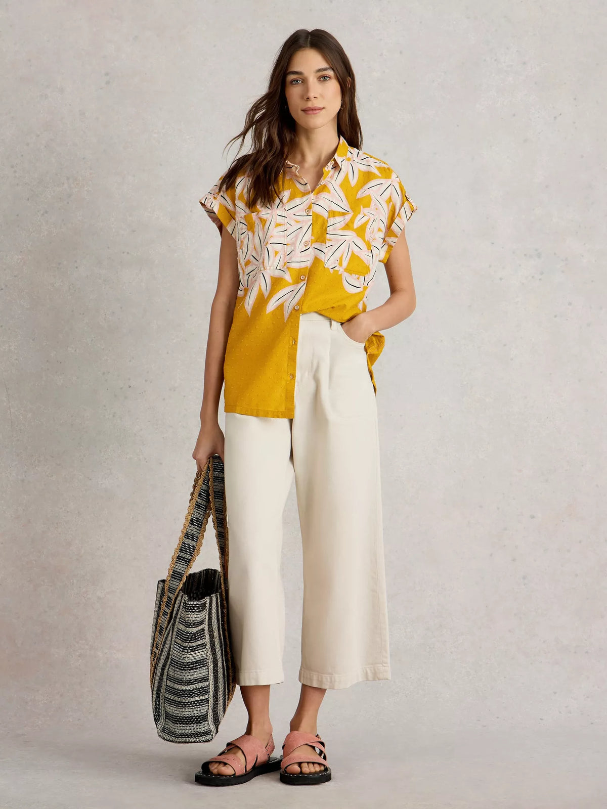 Woman wearing a yellow and white floral blouse with white pants, holding a black and white bag on a light gray background.