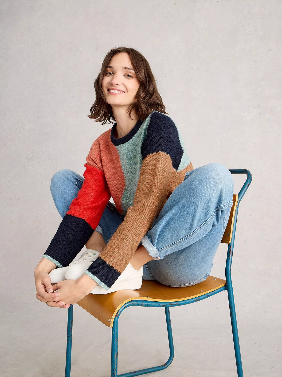 Person sitting on a chair wearing a colorful sweater and jeans against a plain background