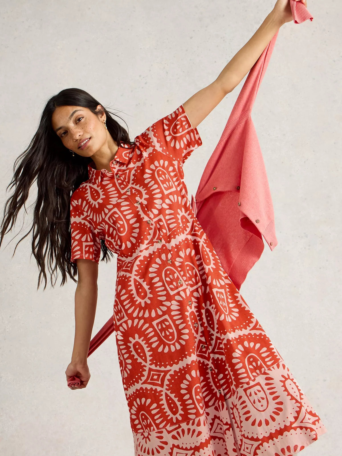 Woman wearing a red and white patterned dress holding a pink scarf against a neutral background