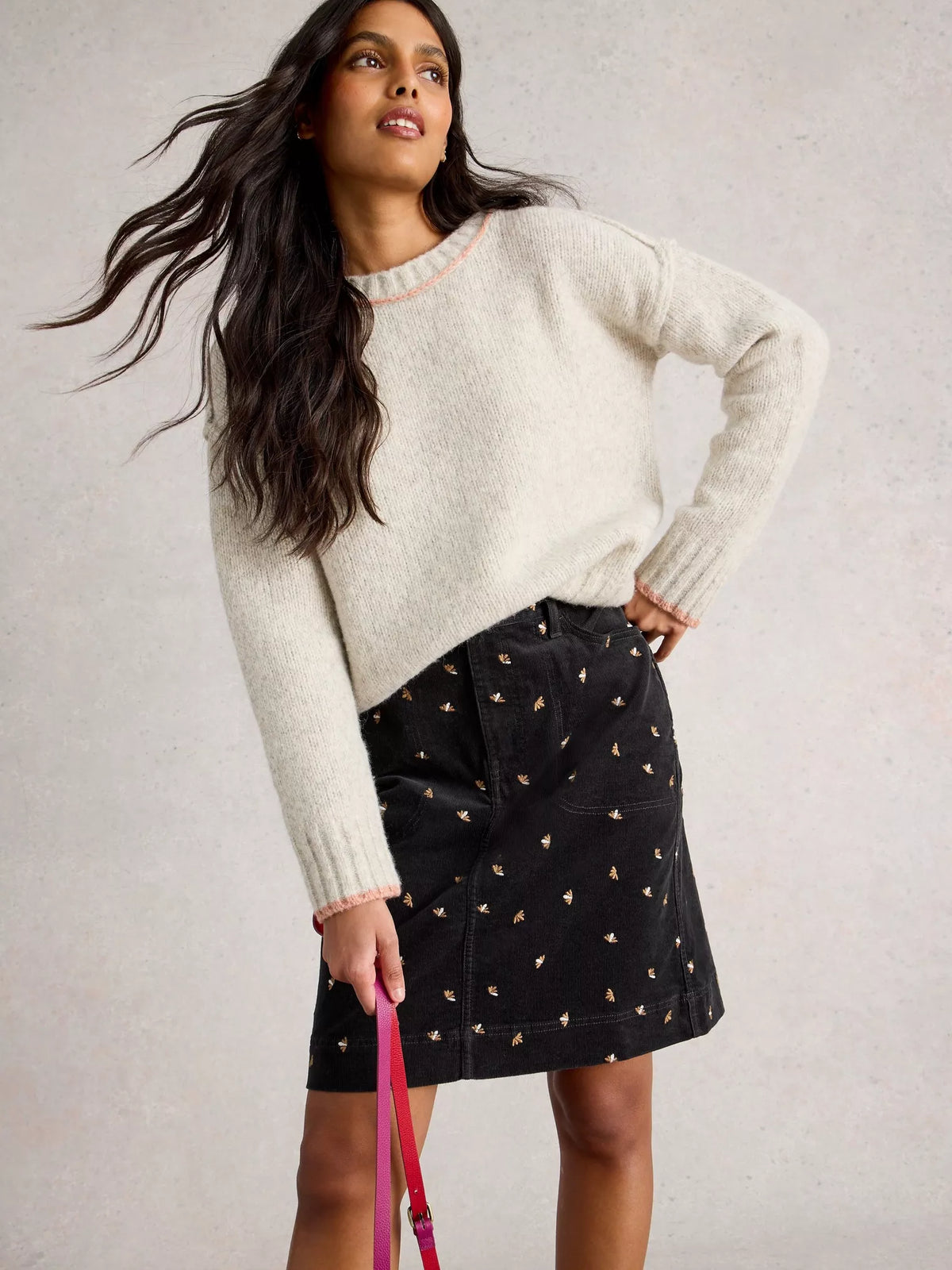 Woman wearing a white sweater and black skirt with gold embellishments on a light gray background