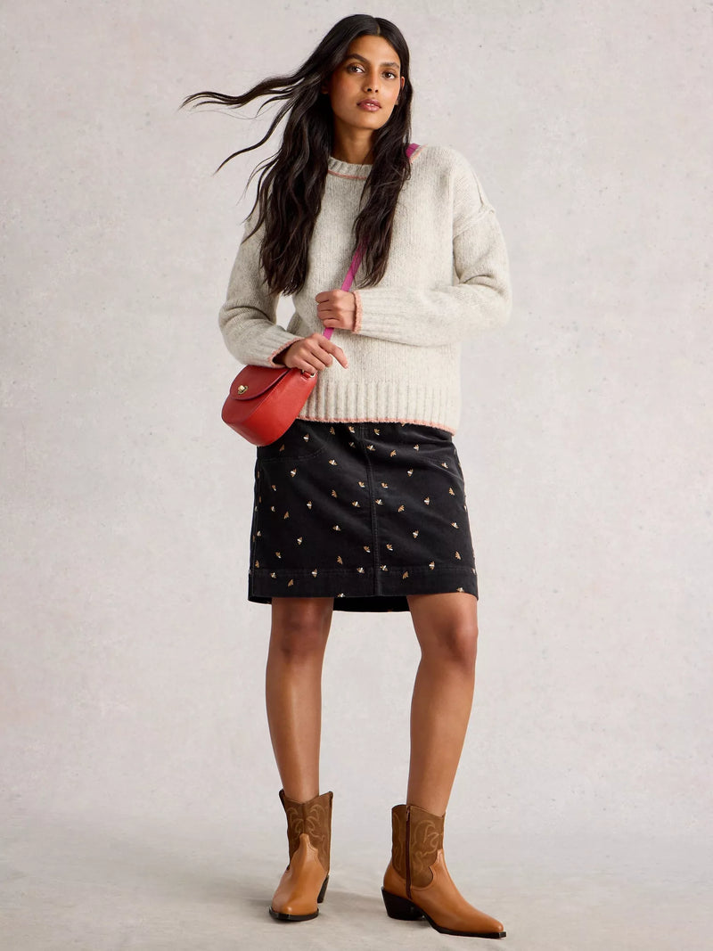 Woman wearing a white sweater, black skirt with gold patterns, and brown boots, holding a red handbag.