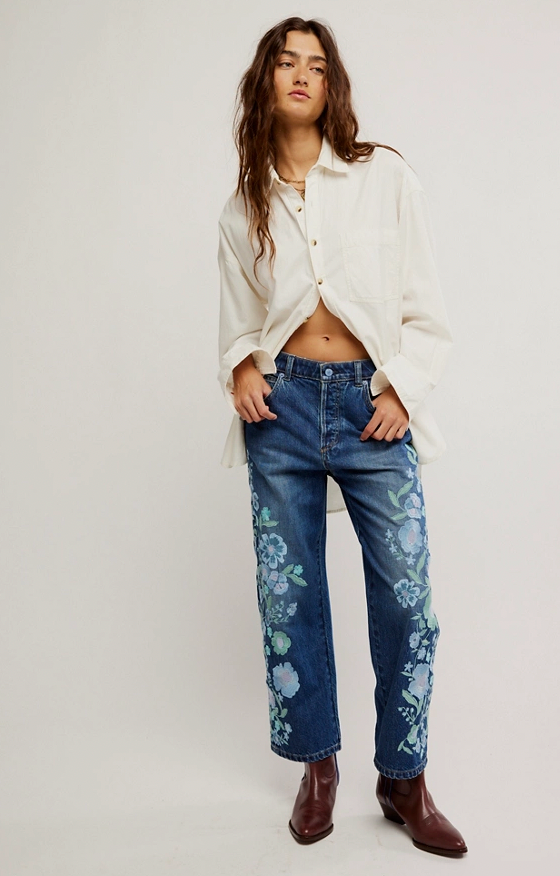 Woman wearing a white shirt and blue jeans with floral patterns on a plain background