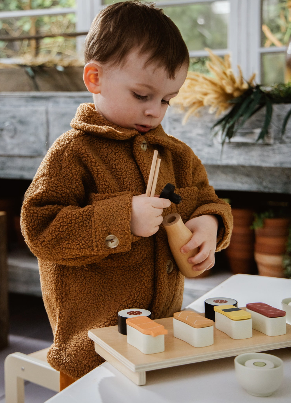 Child in a brown coat holding chopsticks over a display of miniature sushi.
